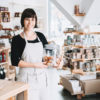 Small local business owner. Seller assistant with glass jar of pastries in interior of zero waste shop. Cheerful woman in apron stands behind counter with food products in plastic free grocery store.